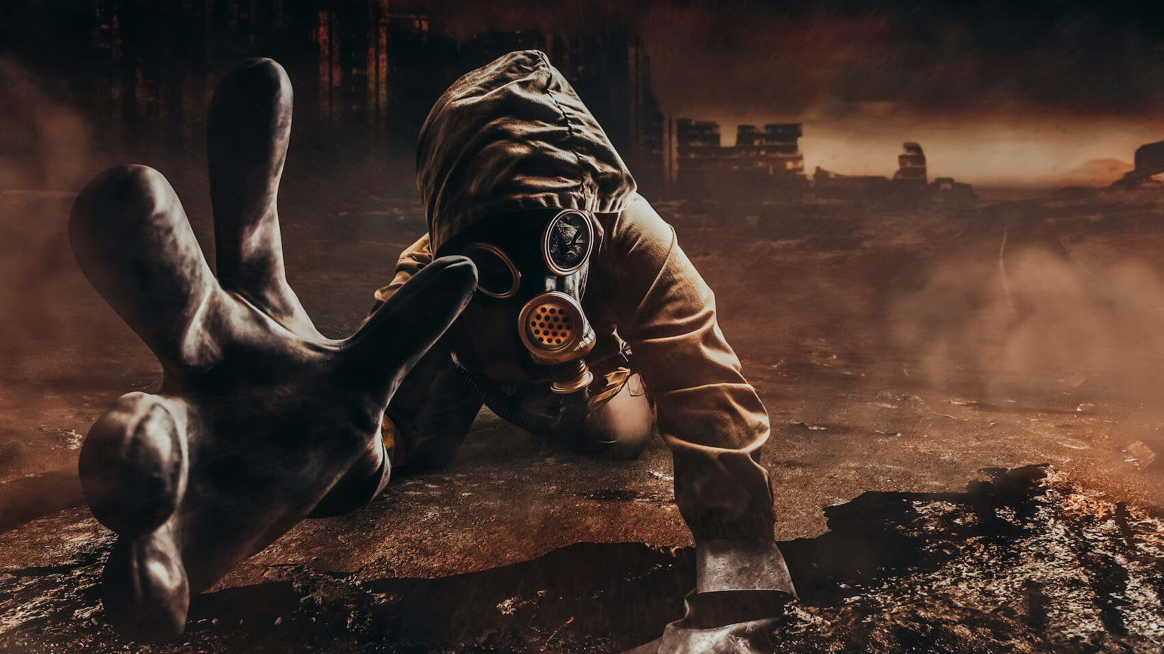 A dying stalker in jacket and gloves in damaged gas mask with filter reaching out his hand to camera on destructed apocalyptic wasteland city background.