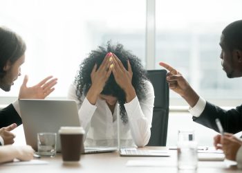 A distressed Black woman professional faces gender discrimination and workplace bullying, as male colleagues point fingers and place blame. The image highlights the emotional toll of inequality and harassment in professional environments. Photo: Dreamstime.