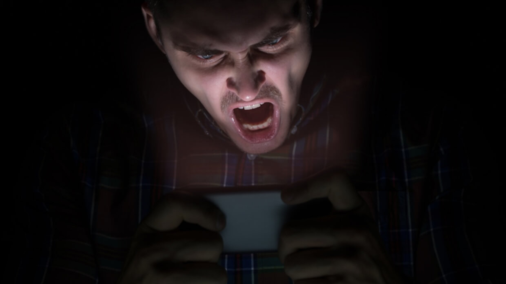 A man sits in the dark, staring angrily at his mobile phone. Photo: Raman Mistsechka.