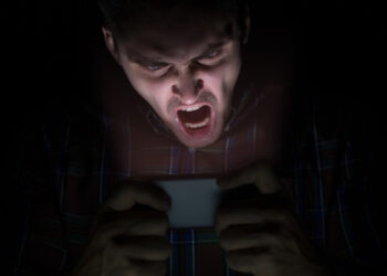 A man sits in the dark, staring angrily at his mobile phone. Photo: Raman Mistsechka.